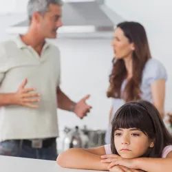 A saddened child sits at a table while parents argue behind her